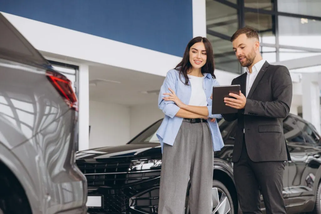 Junge Frau berät sich mit einem Verkäufer und hält ein Tablet, während sie ein Auto in einem Autohaus auswählt, um ein neues Fahrzeug zu kaufen.